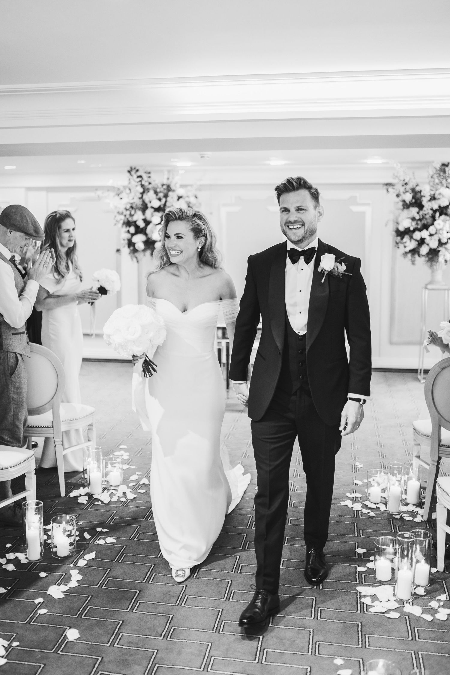 A black and white wedding photo of a couple walking down the aisle with floral arrangements and scattered petals.Claridge’s wedding