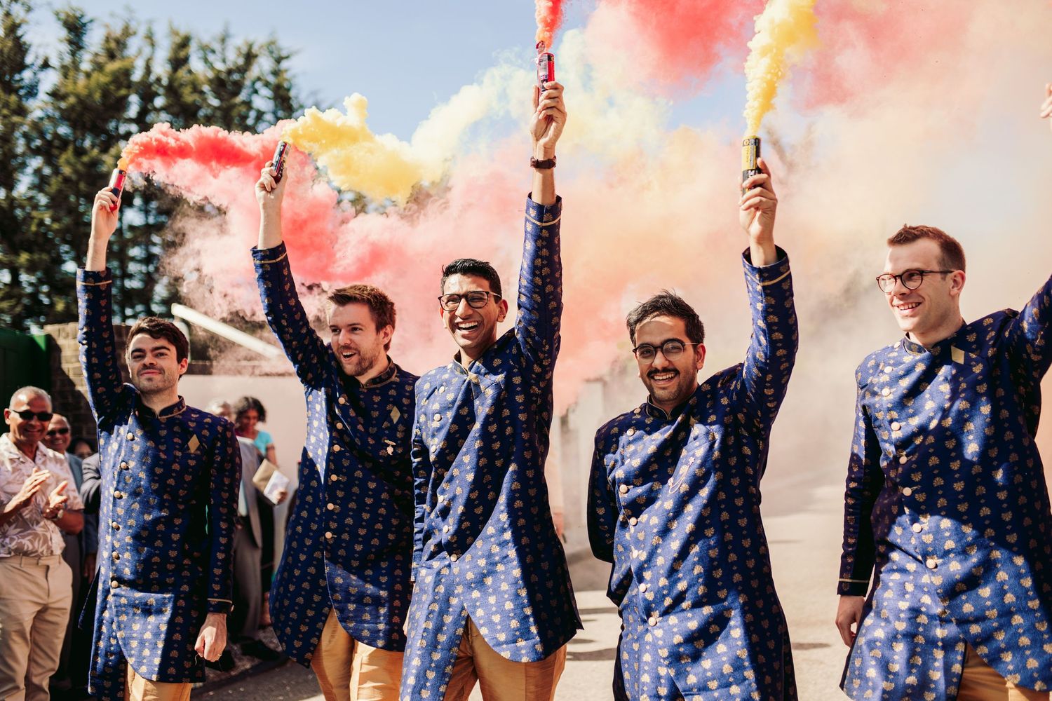 A group of groomsmen in matching patterned suits hold colorful smoke bombs during a wedding celebration outdoors.