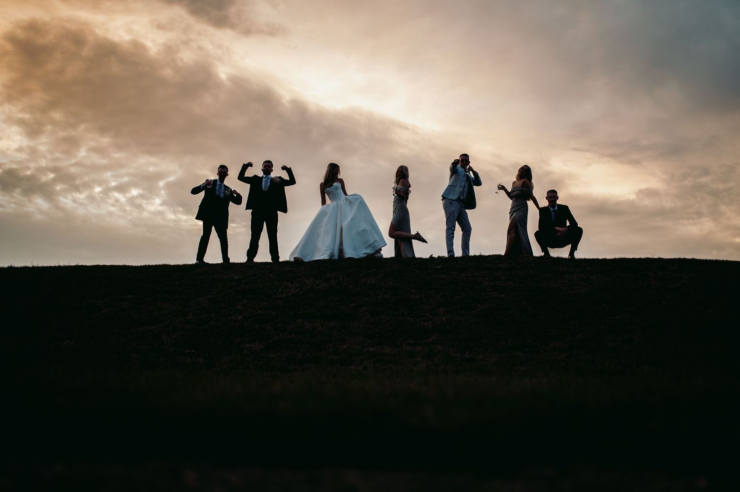Silhouettes of wedding party posing on hill against dramatic sunset sky.