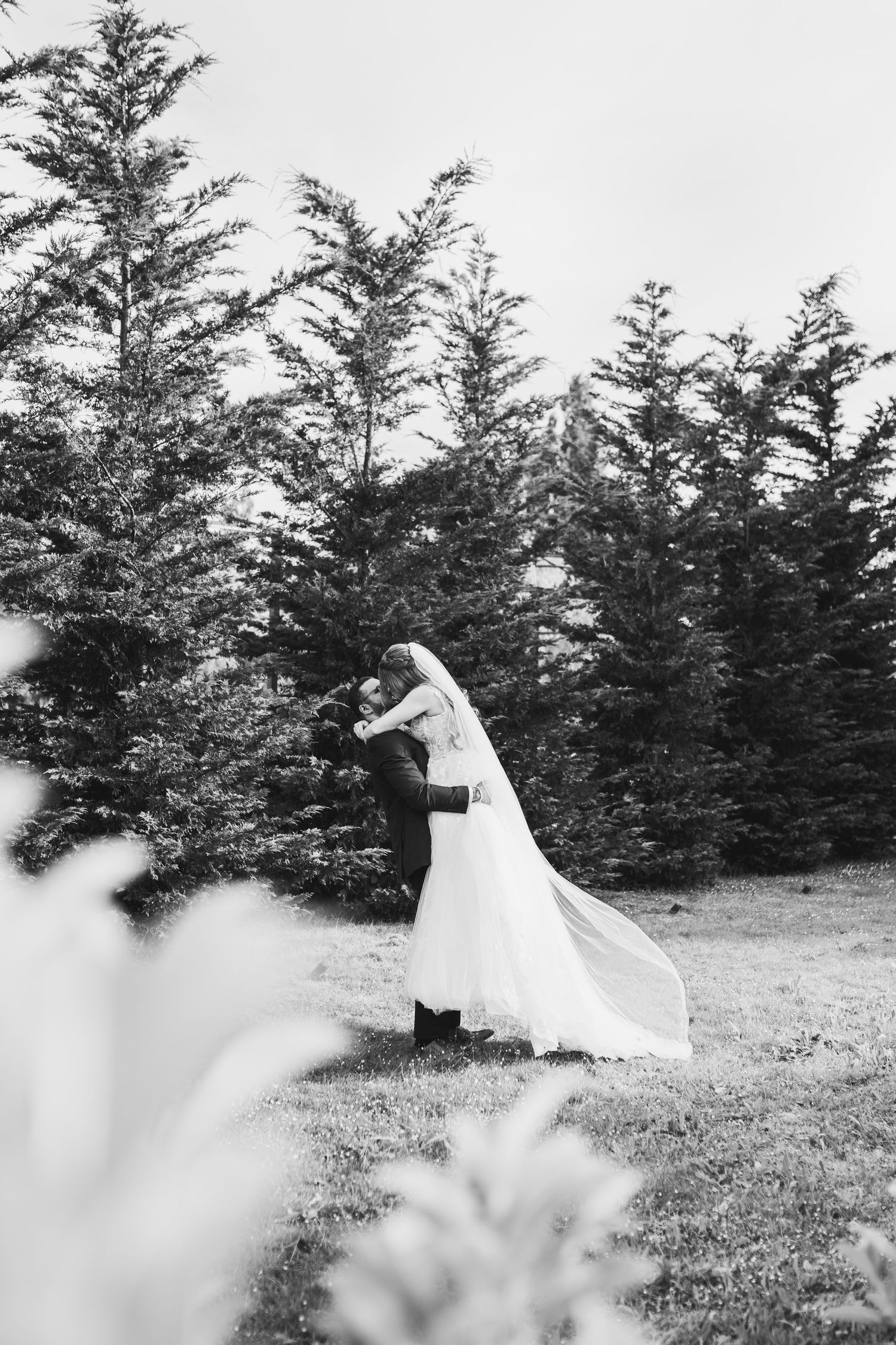 Black and white wedding photo showing couple embracing in a forest clearing with tall evergreen trees in background.