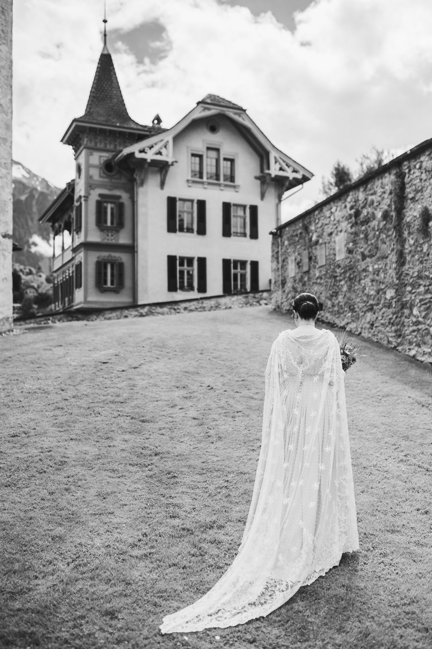 A figure in a flowing white cape stands on a path in front of a Swiss chalet-style house with a tower spire. Spiez switzerland destination