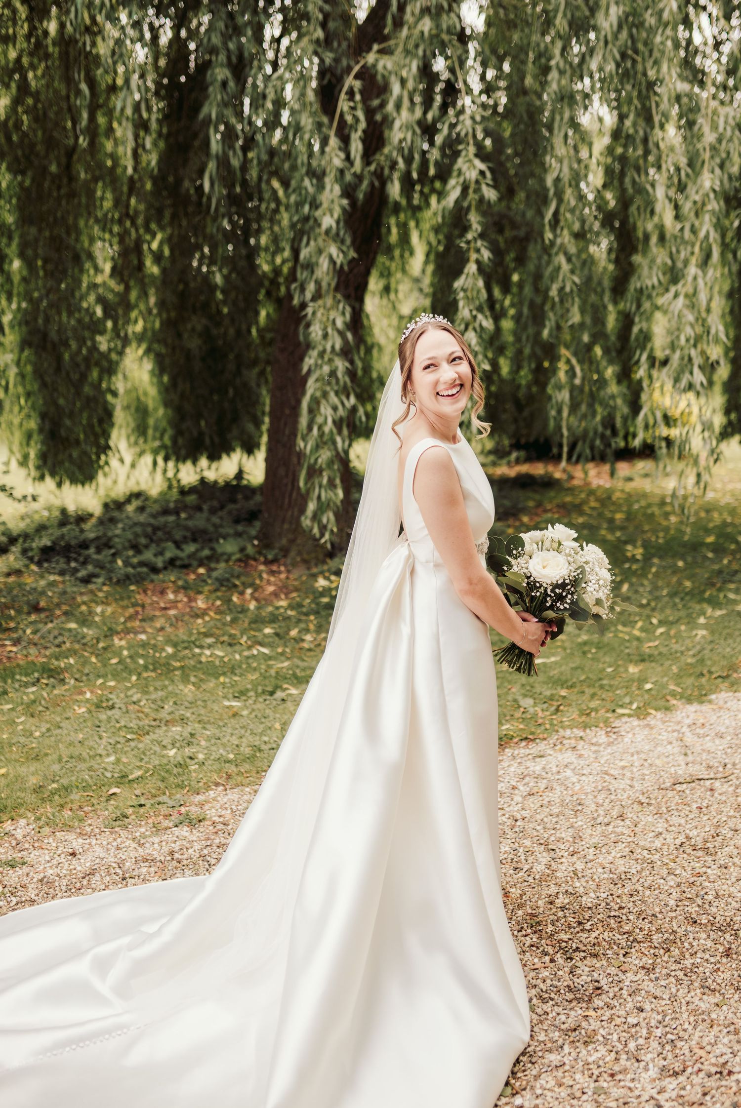 A bride in a flowing white wedding dress poses gracefully under weeping willow trees on a gravel path.