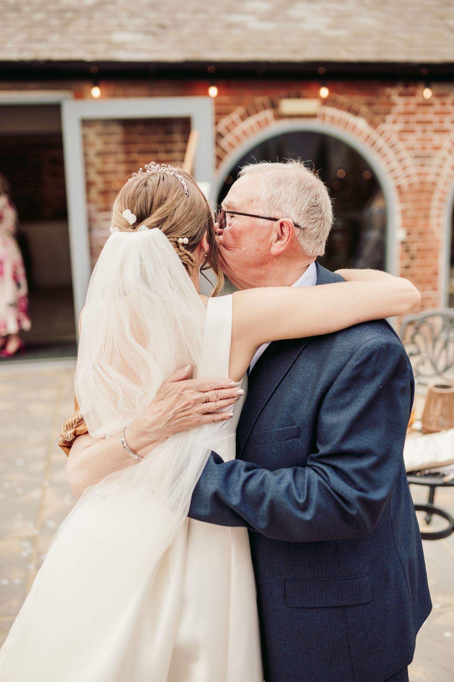 A touching moment captured at a wedding with a navy suit and white dress against a brick wall backdrop.