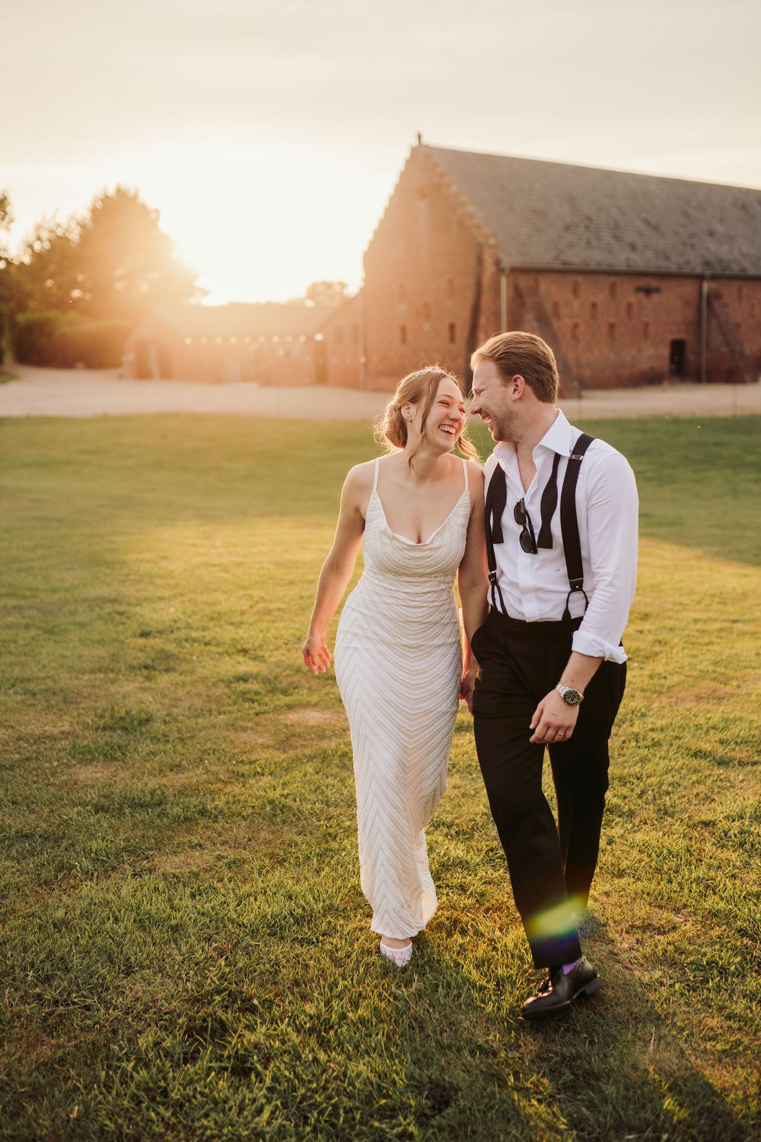 A couple in wedding attire walks together through a sunlit grassy field during golden hour.