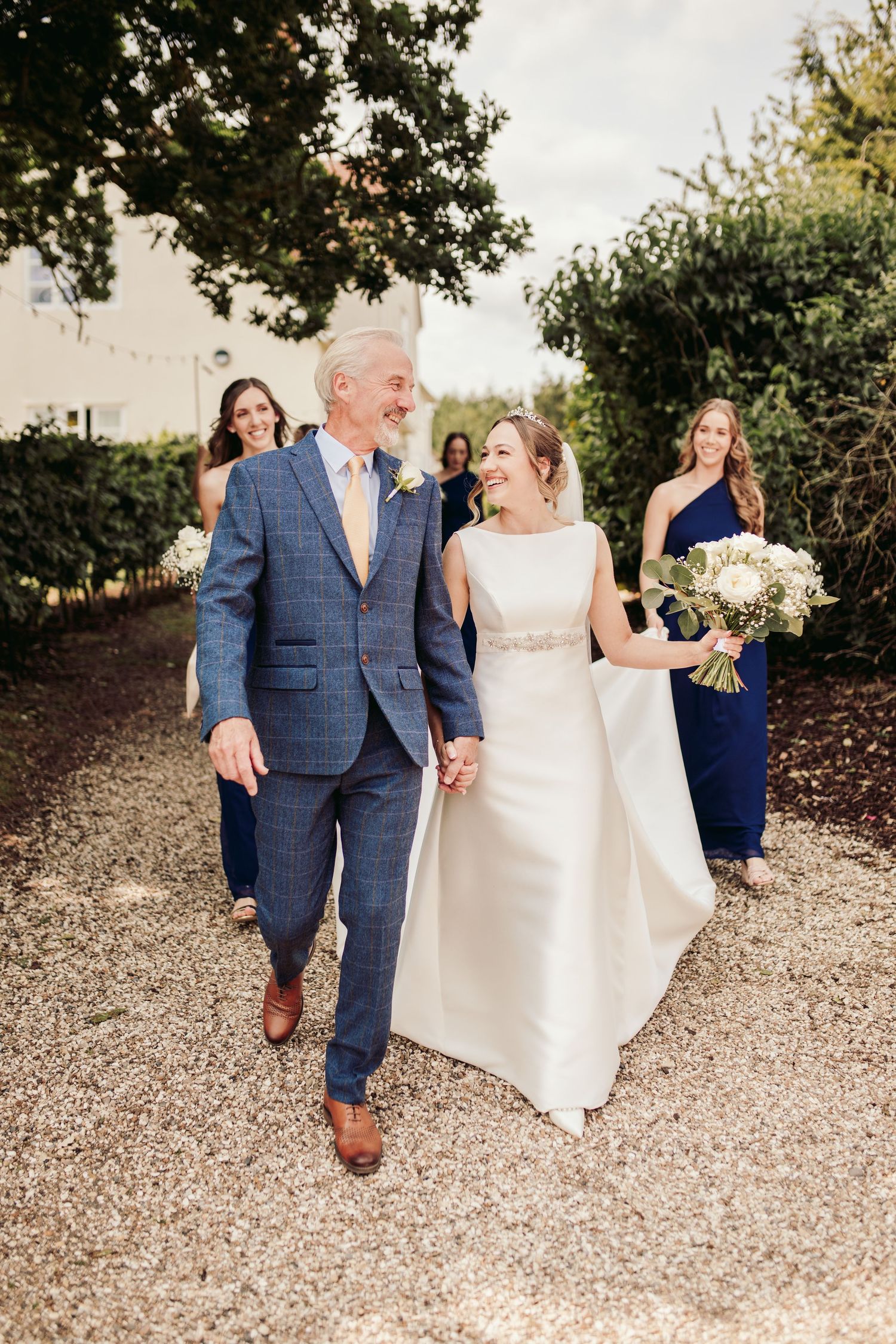 A couple walks together on a gravel path surrounded by greenery during a sunny outdoor celebration.