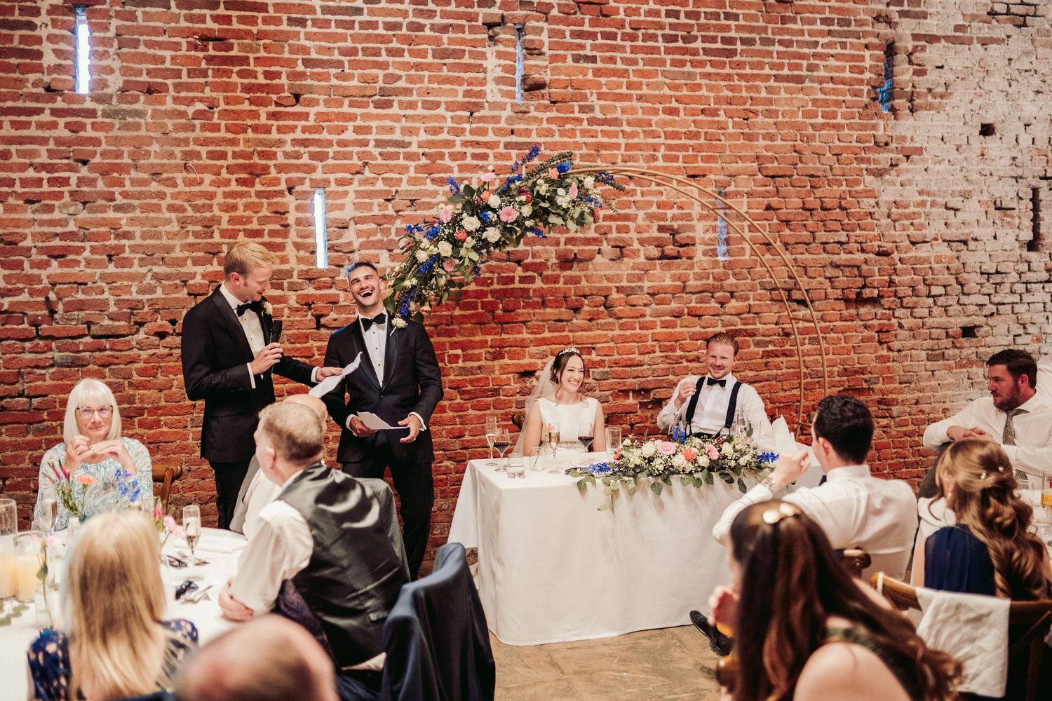 Wedding reception guests seated at tables in a rustic brick-walled venue with string lights and floral decorations.