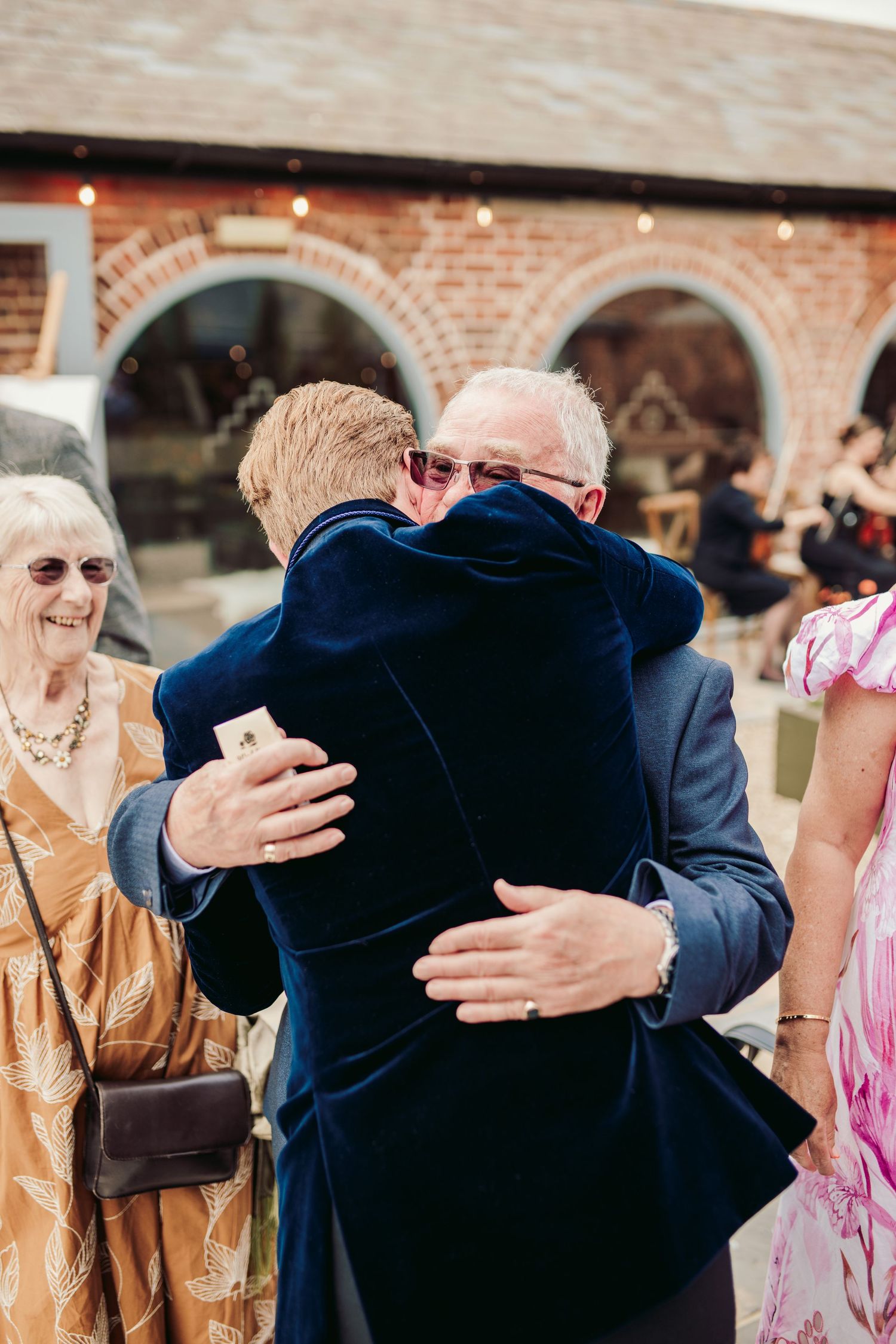 Two people share a warm embrace at an outdoor event with brick archways visible in the background.