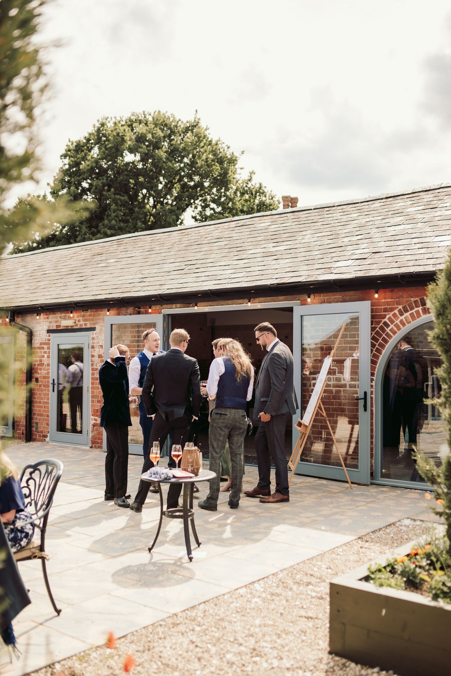 A group gathers on a stone patio outside a brick building with glass doors and a slate roof on an overcast day.