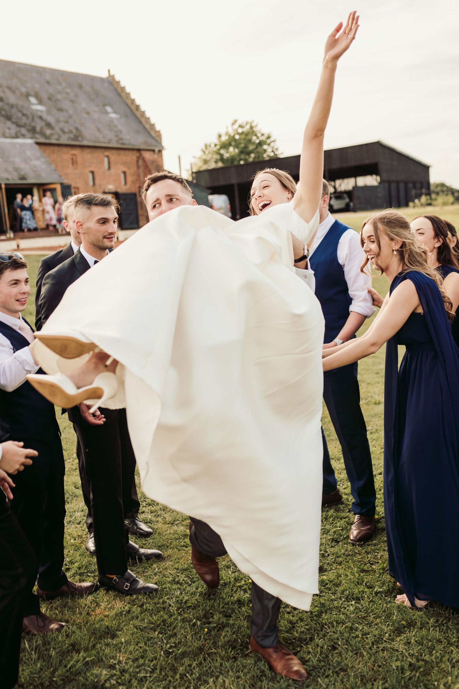 Wedding party in navy and white attire celebrates joyfully on grassy lawn outside rustic brick building.