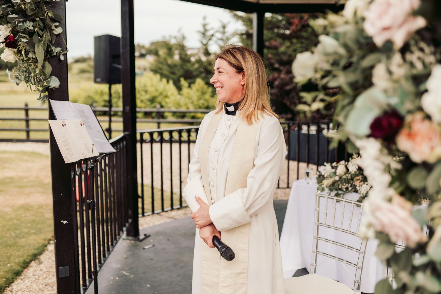 A woman in a white dress officiates an outdoor wedding ceremony with floral decorations at a garden venue.