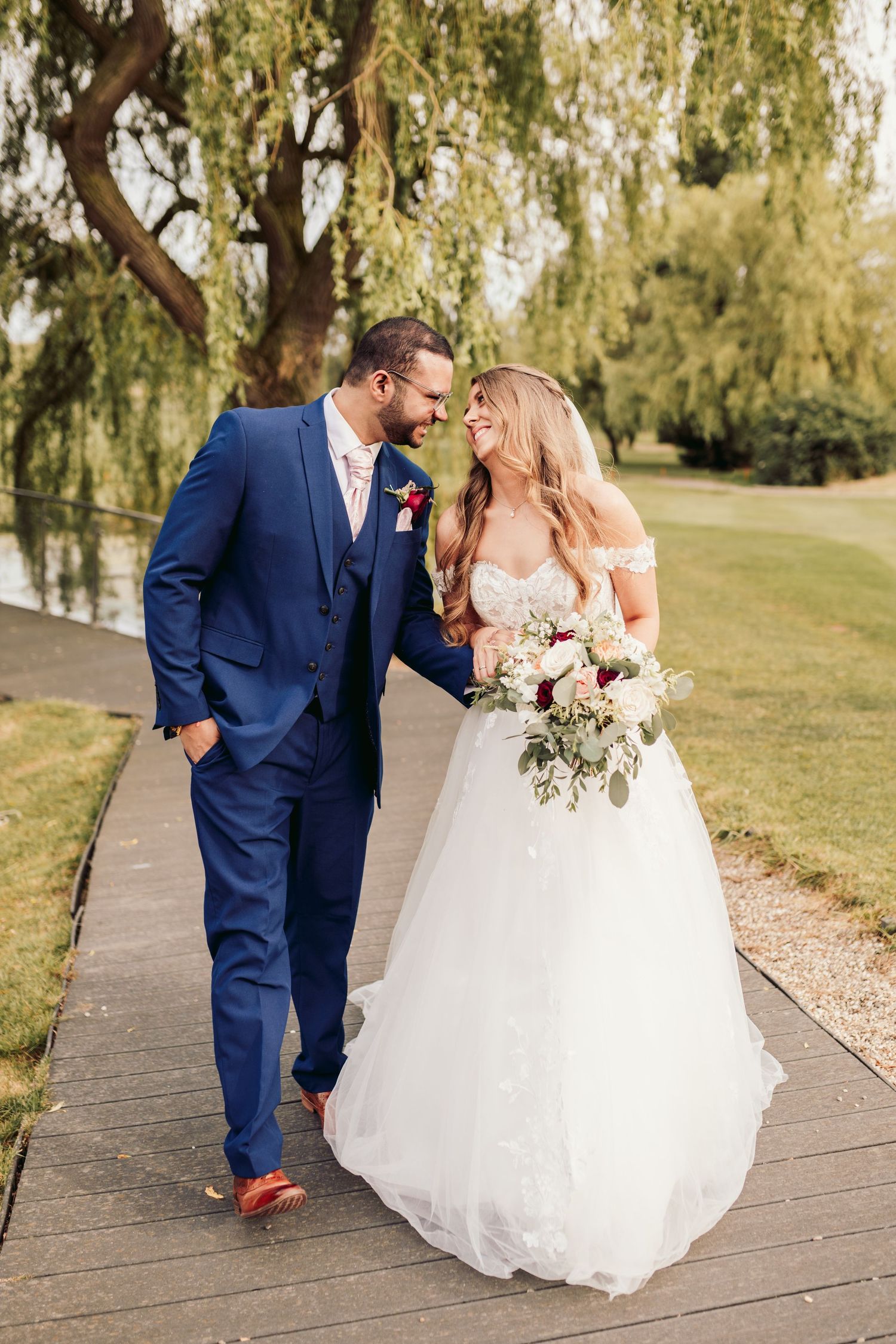 Newlywed couple walks together on wooden pathway under willow tree wearing blue suit and white wedding gown.