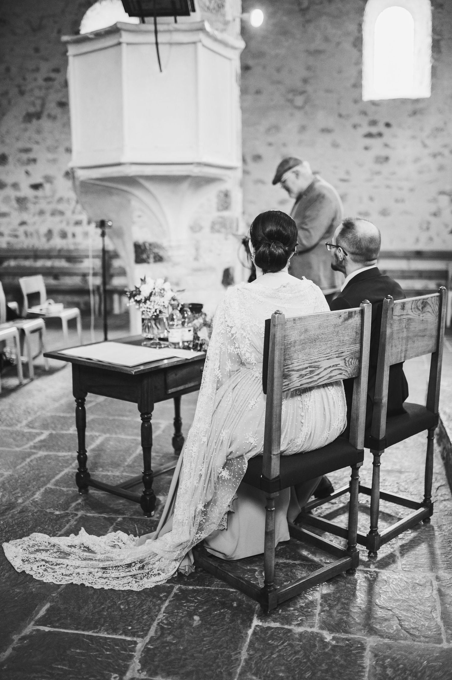 Black and white photo of couples seated at a wedding ceremony in church with white pulpit and stone walls. Spiez switzerland destination