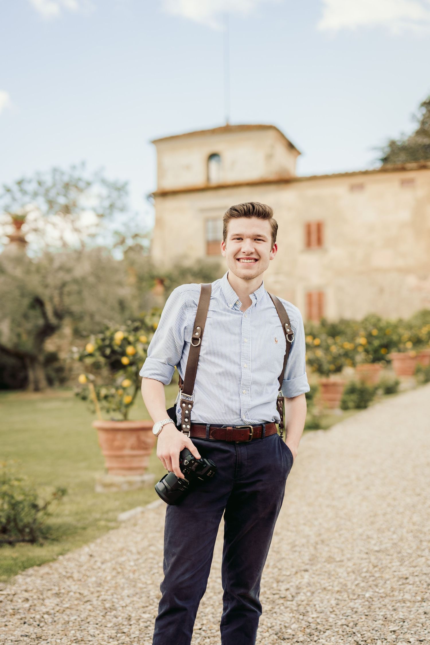 Wedding photographer wearing suspenders and dress shirt poses outdoors at rustic venue.