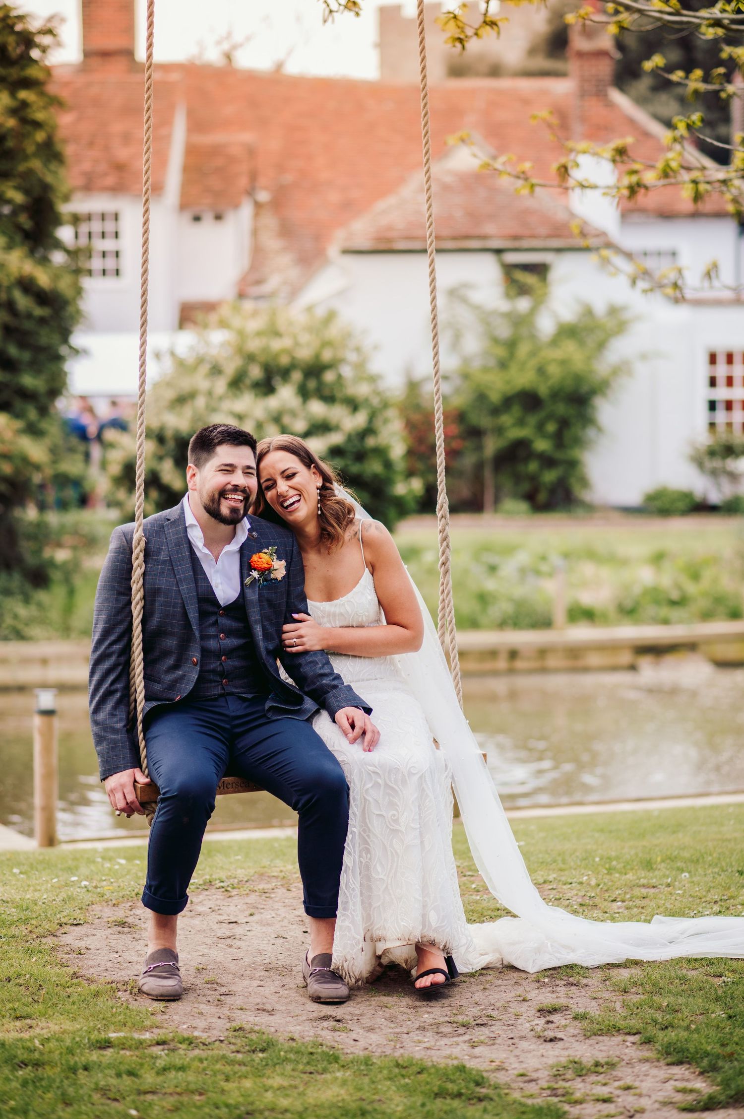 A relaxed wedding couple sits together on a swing by a pond, sharing a happy moment.