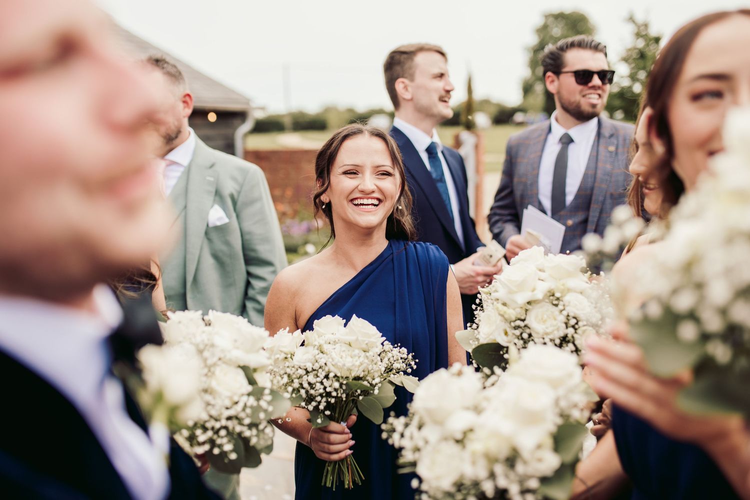 Wedding party laughs together while holding white and green floral bouquets in outdoor garden setting.