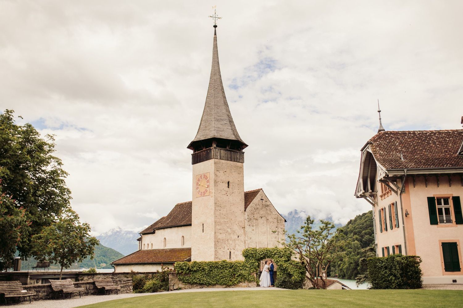 A scenic church with tall spire stands on a hillside in Switzerland surrounded by traditional buildings and green lawns. Spiez switzerland