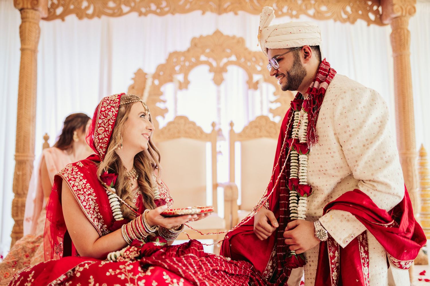 A traditional Indian wedding ceremony shows the couple seated together in ornate red and cream colored wedding attire.