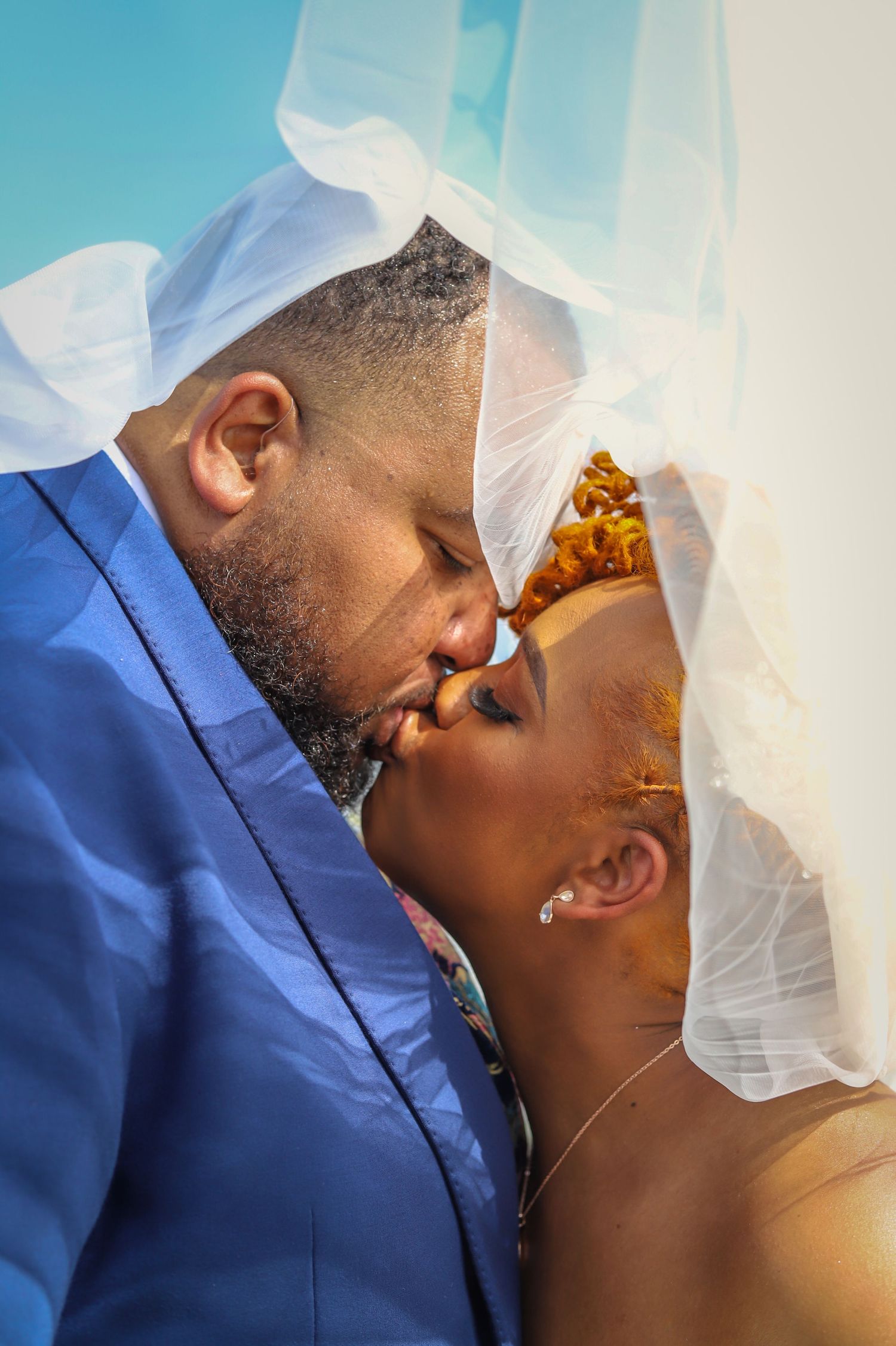 Couple kissing under white veil, groom in blue suit, bride in traditional headwrap.