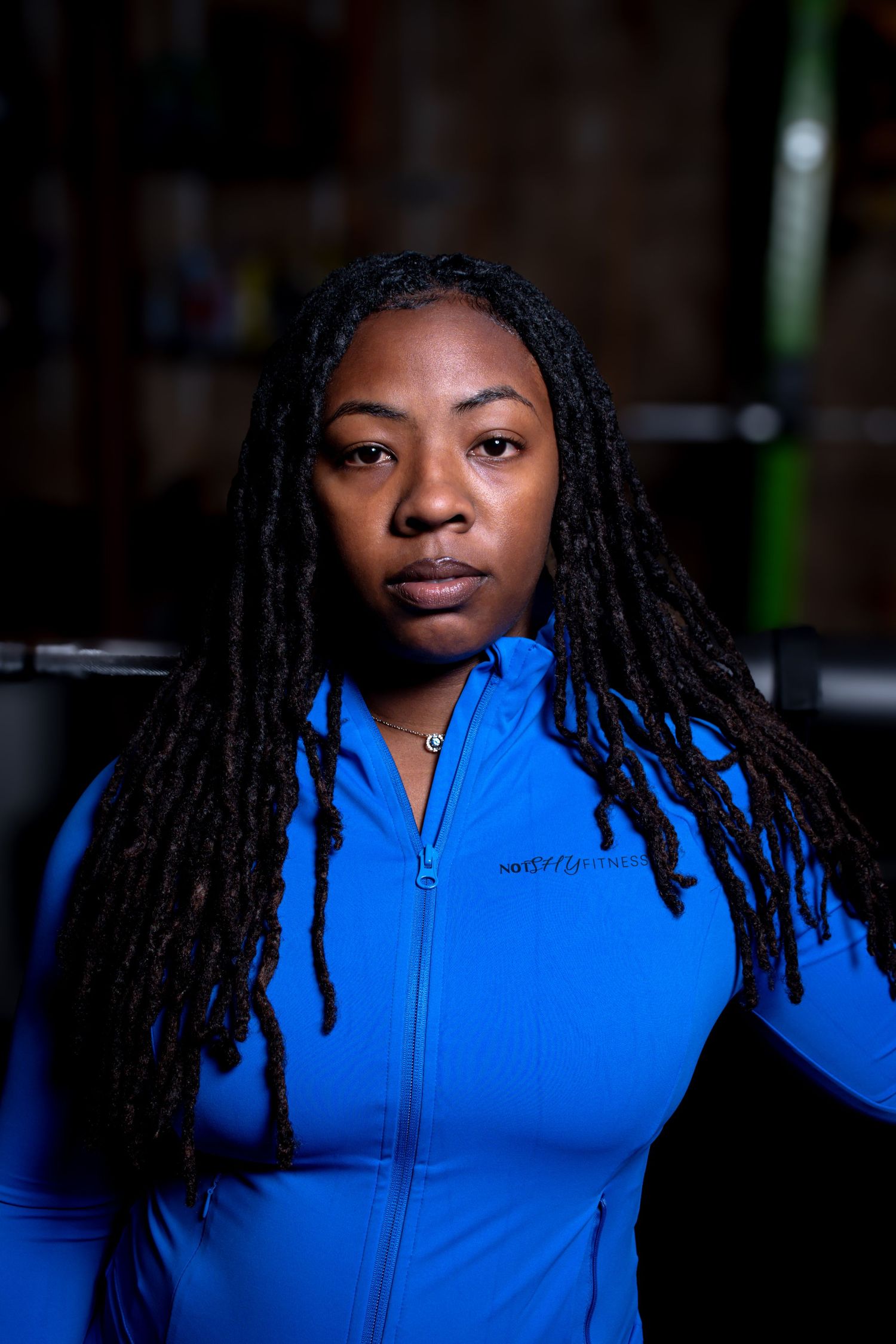 Woman with locs in blue athletic jacket poses seriously in dark gym setting.