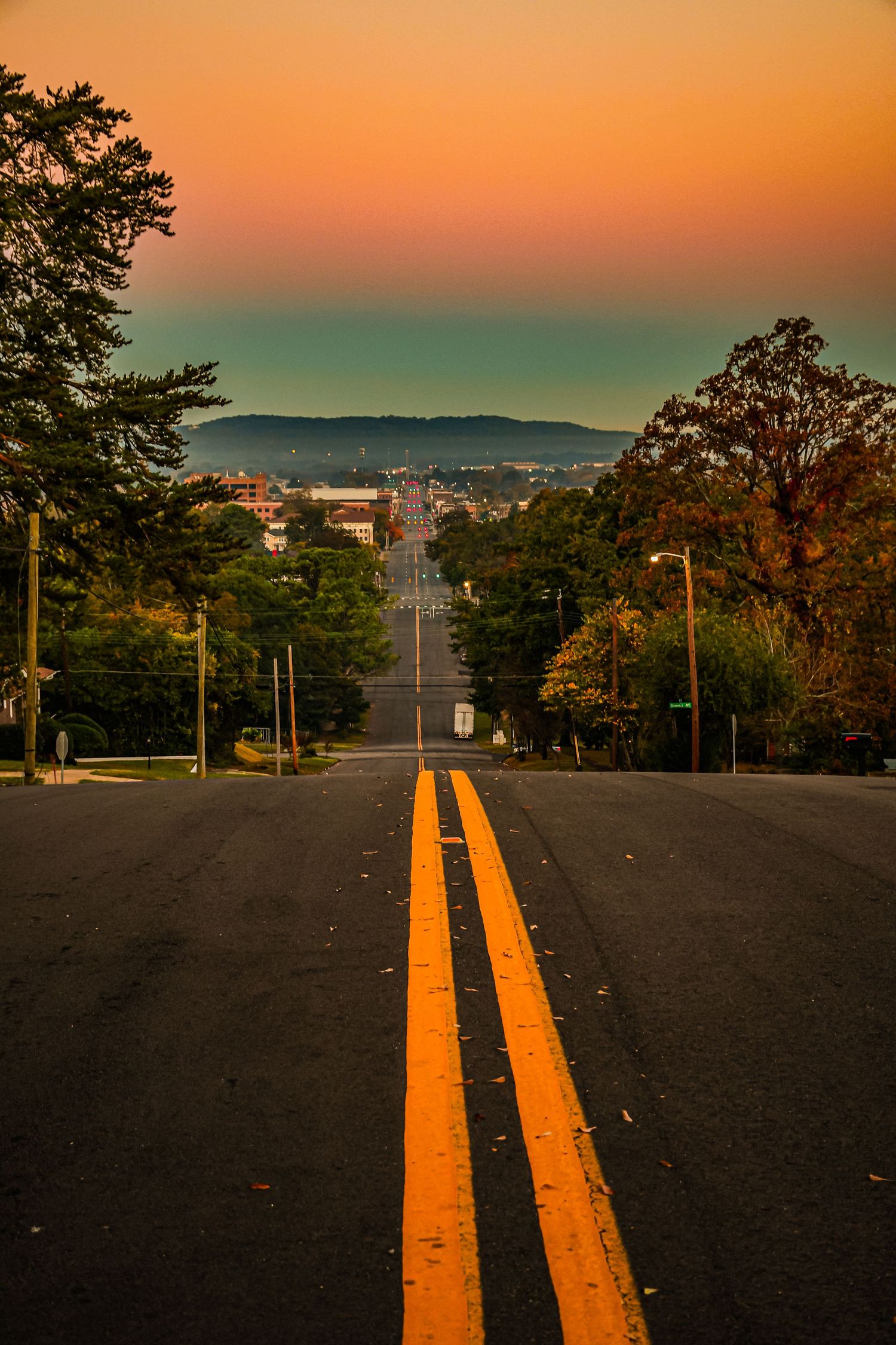 A long empty road with double yellow lines leads into a small town under a vibrant orange and teal sunset sky.
