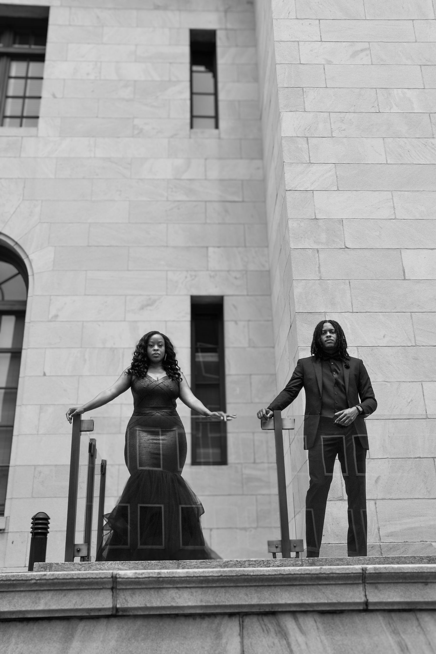 Black and white photo of an elegant couple in black formal wear posing on steps of a stone building.