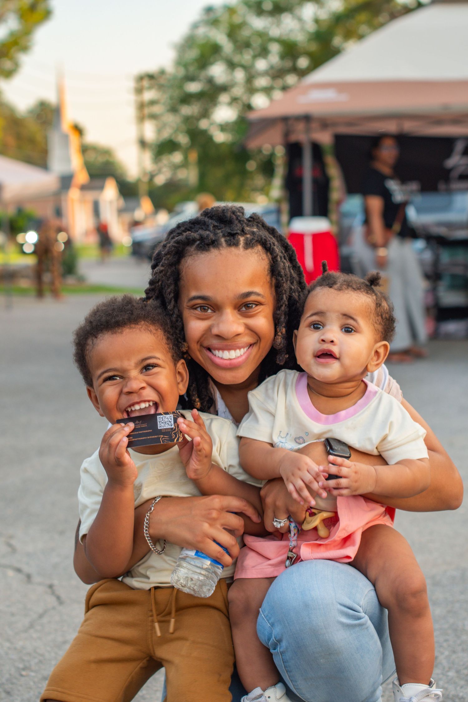 A smiling young woman outdoors holds two happy toddlers, one boy and one girl, at a casual outdoor event.