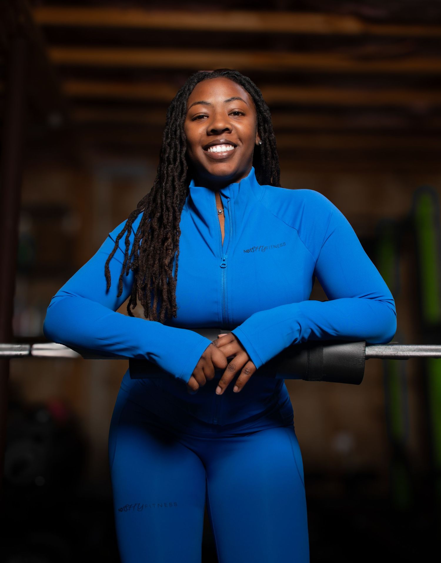 Smiling woman in blue athletic set leans on barbell in rustic gym.