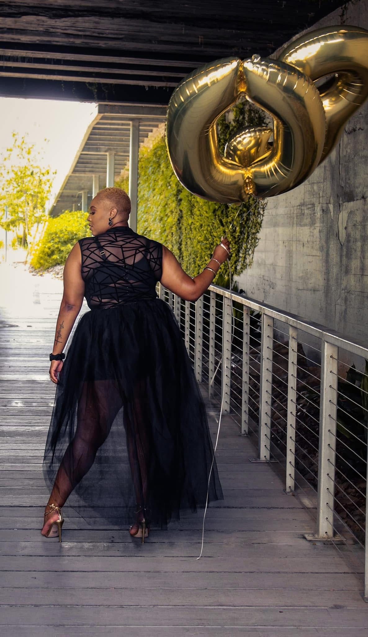 Bald Black woman in black tulle dress holding gold balloons on outdoor walkway.