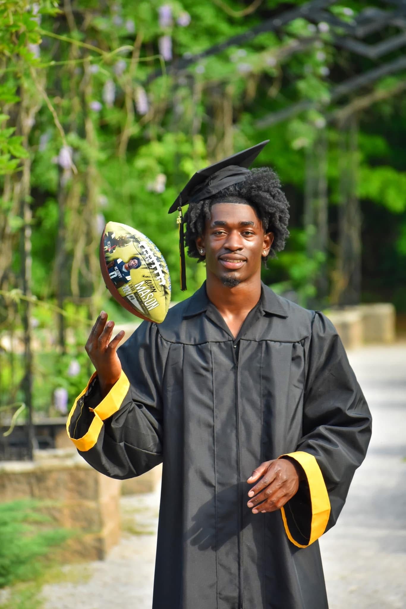 A graduate in cap and gown spins a decorated football outdoors in a green garden.