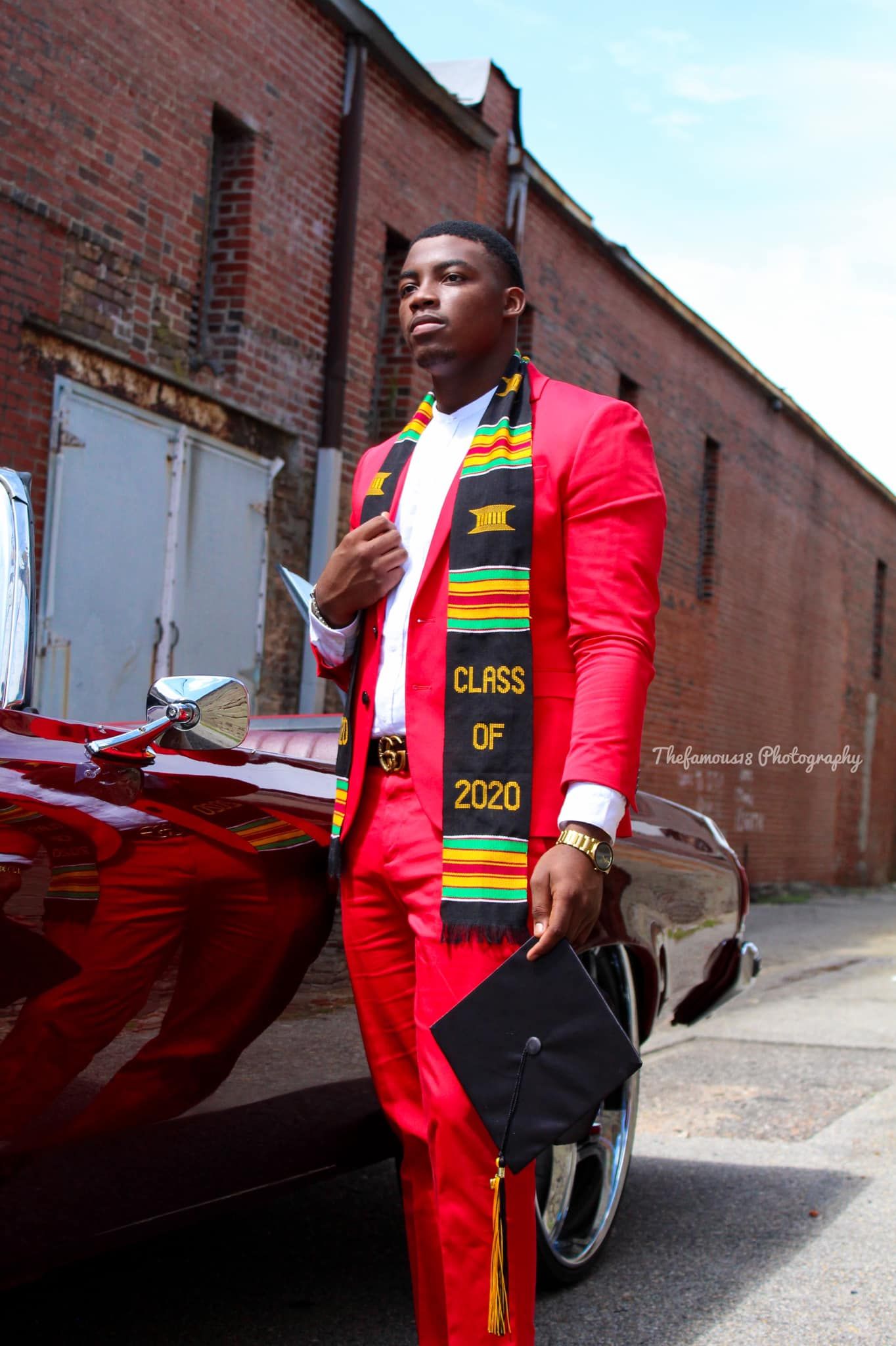 A young Black male graduate in a red suit wearing a Class of 2020 kente stole poses beside a red classic car.