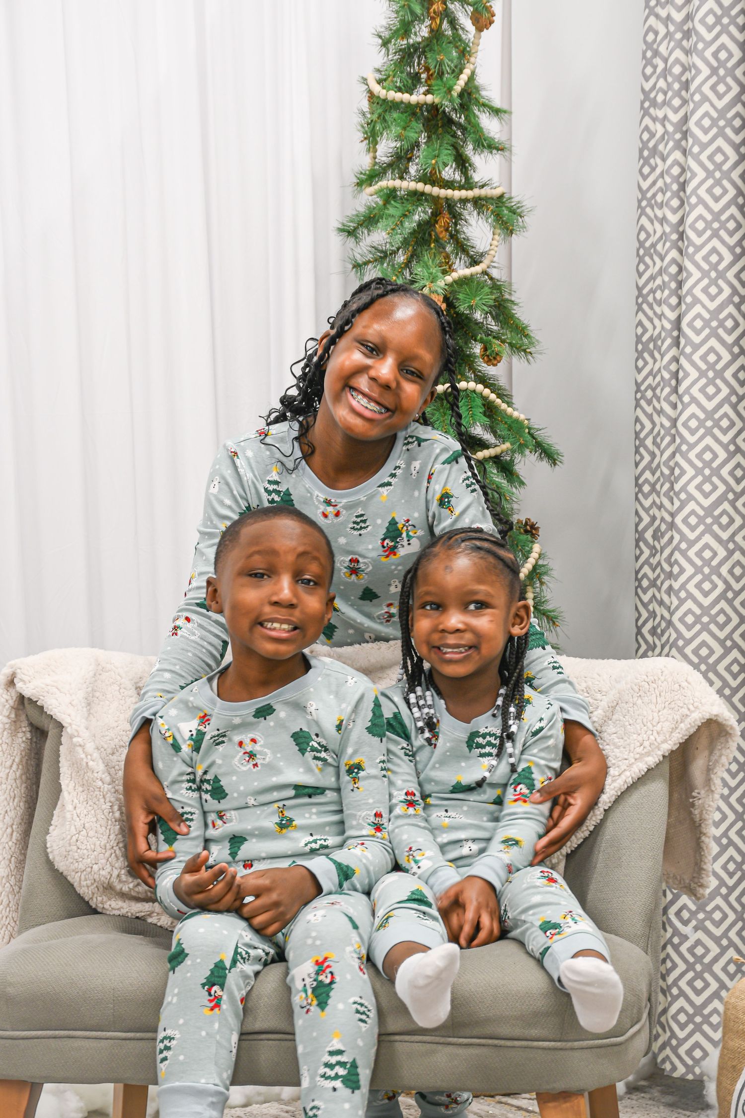 A mother and two young children wear matching Christmas pajamas seated by a decorated tree.