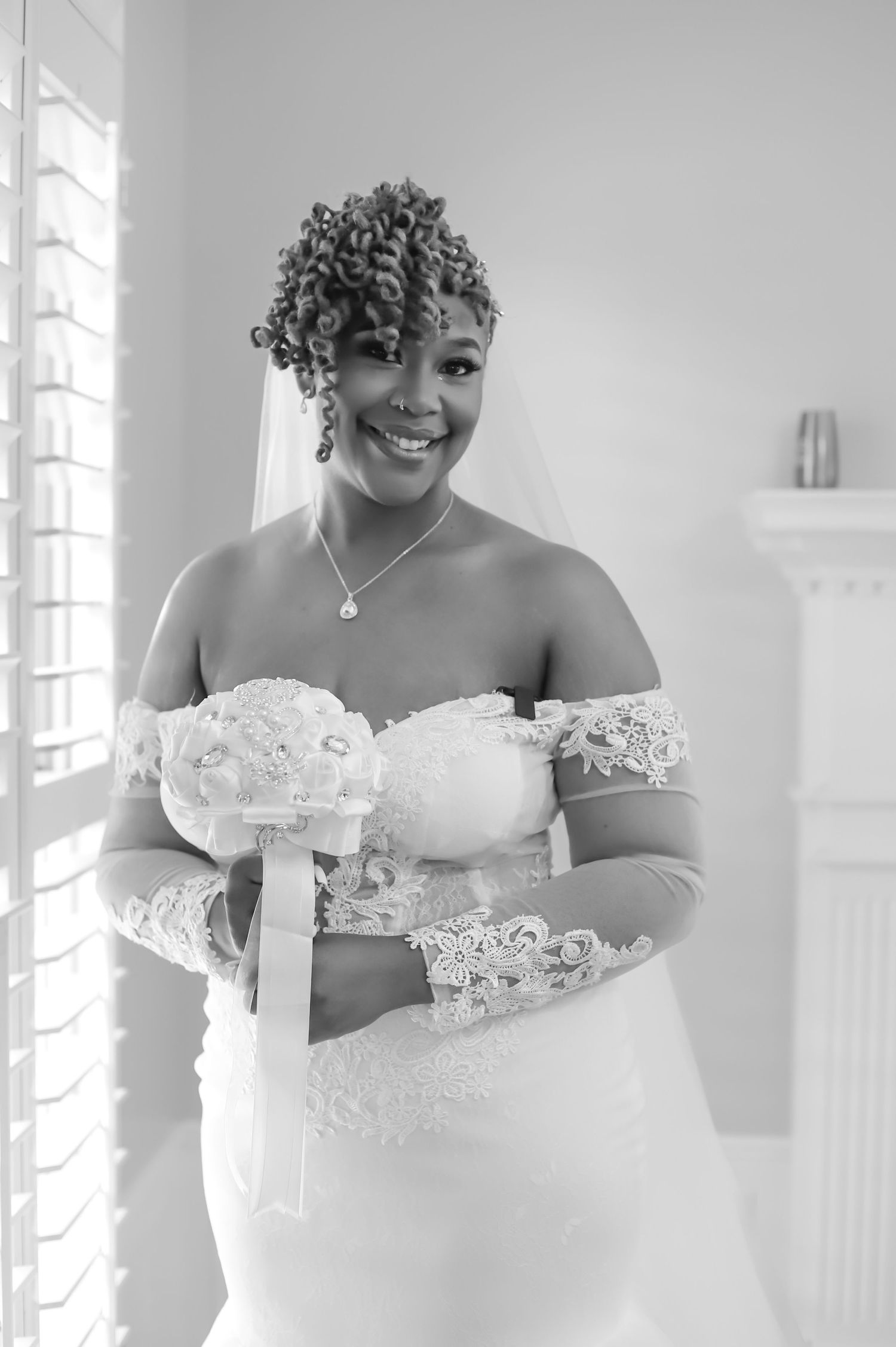 A radiant Black bride in a white lace gown holds a bouquet and smiles in a black and white photo.