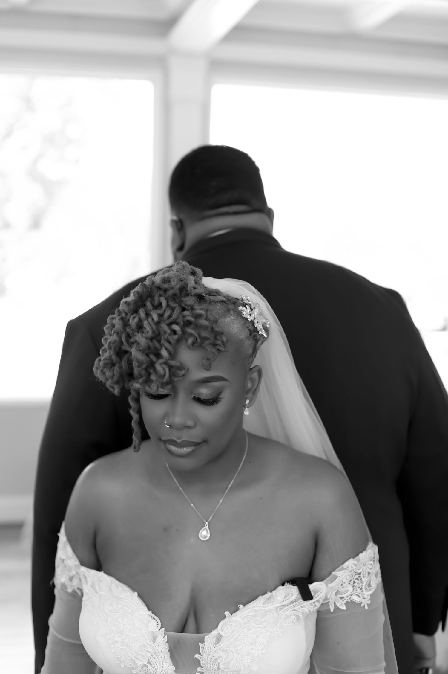 Elegant bride in lace off-shoulder gown with curly updo stands near groom in black and white photo.