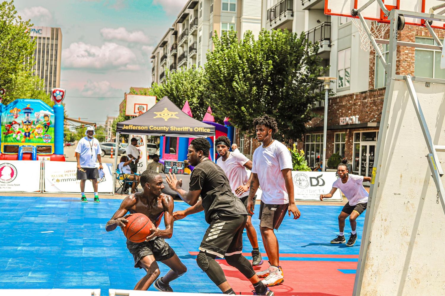 Young athletes compete in an intense outdoor 3-on-3 basketball game on a vibrant blue court.
