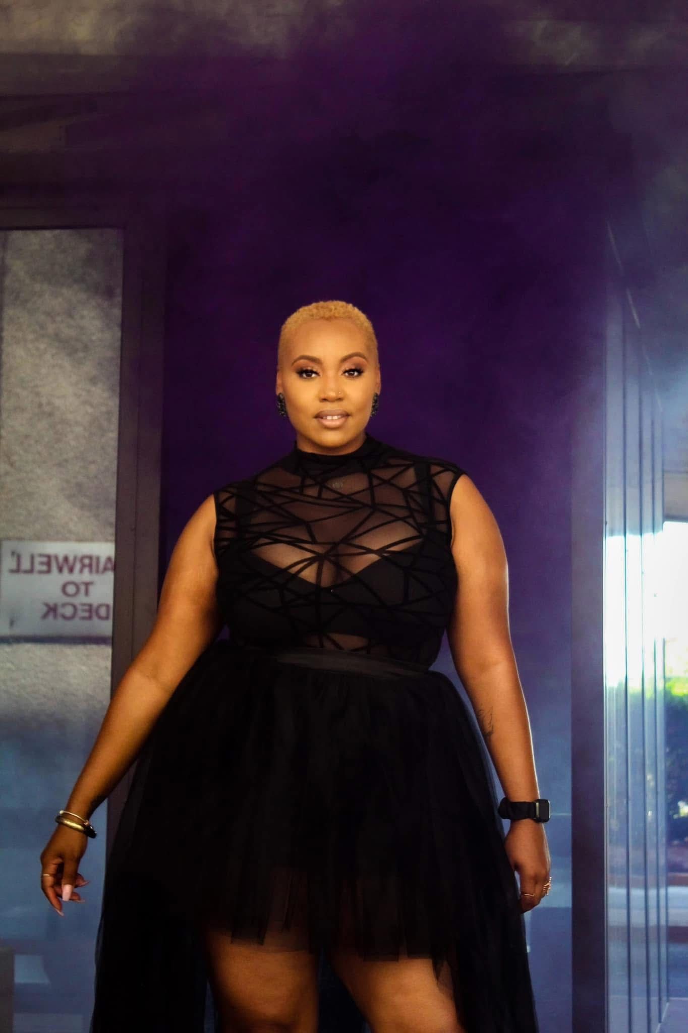 Woman in black sheer dress posing dramatically with purple smoke in background.