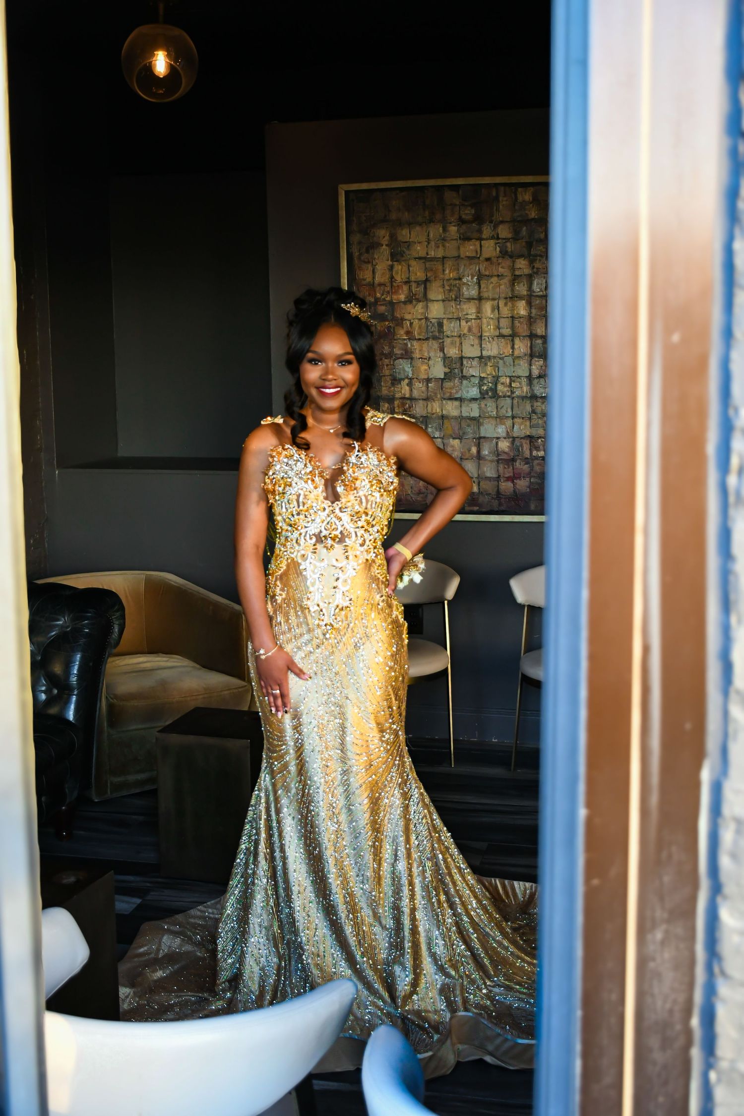 Smiling woman in ornate gold sequin gown stands in elegant darkly decorated room doorway.