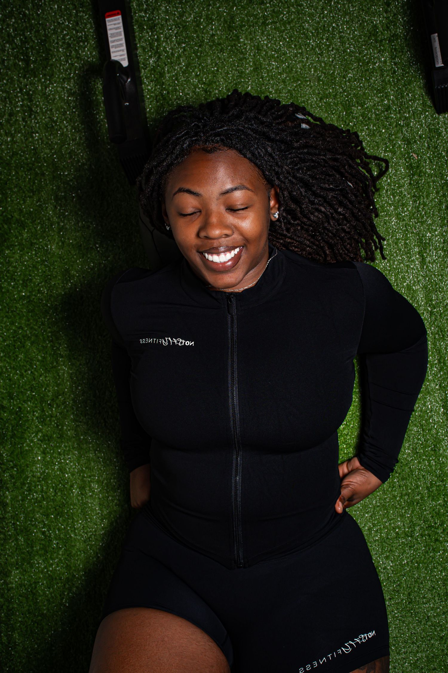 Black woman with locs in black zip-up workout outfit smiling while lying on gym turf.