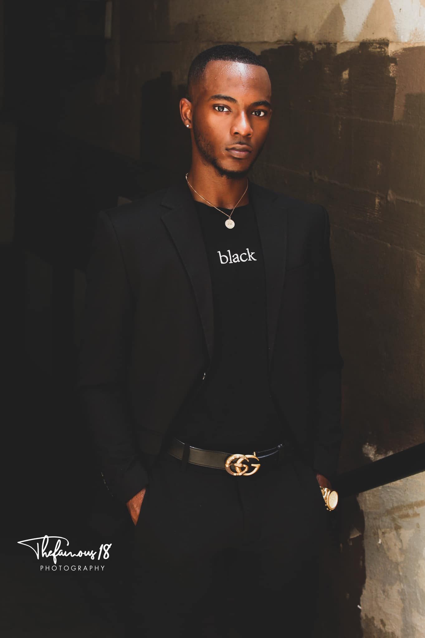 A stylish young Black man in a black suit with Gucci belt poses against a dark textured wall.
