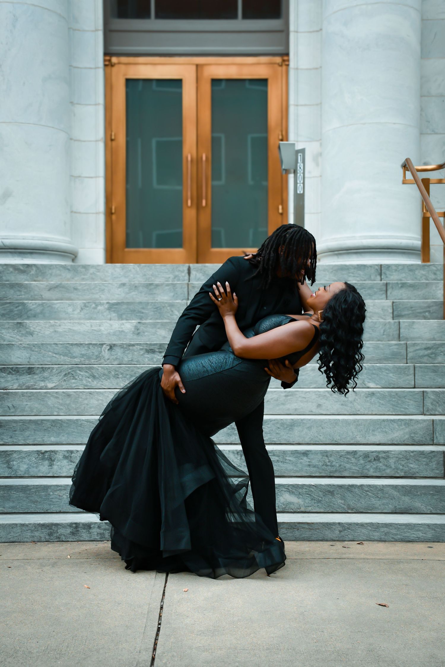 Couple in black formal wear sharing romantic dip pose on marble steps of historic building.