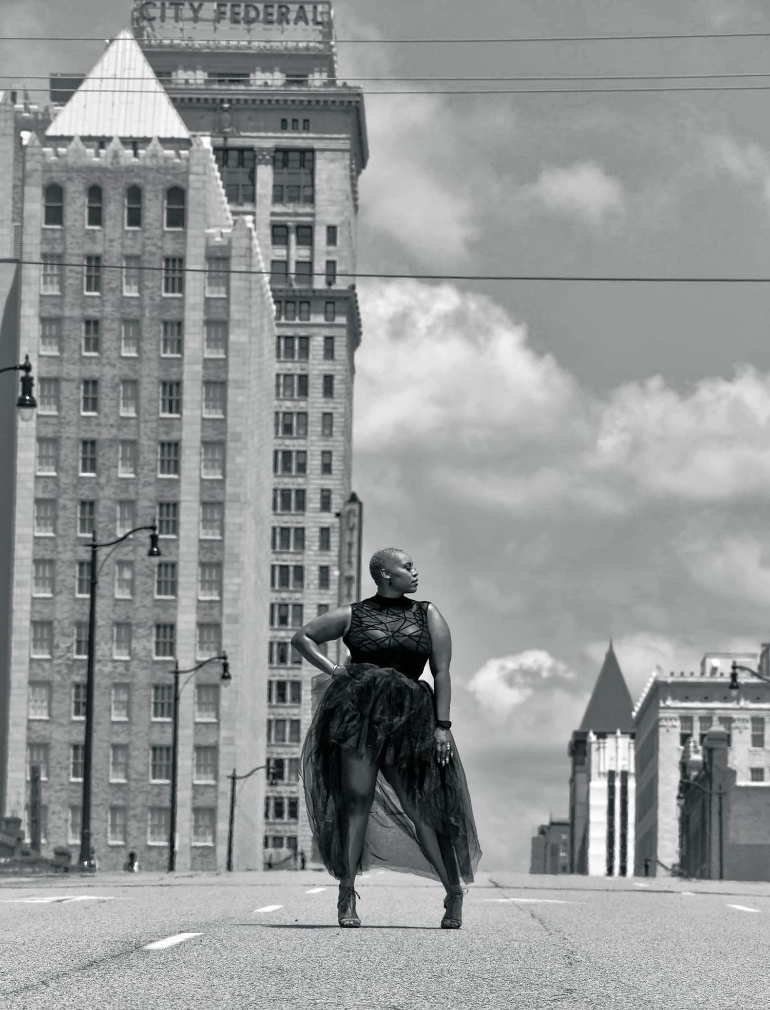 Black and white photo of a woman in a flowing dress posing on an empty city street with tall buildings.