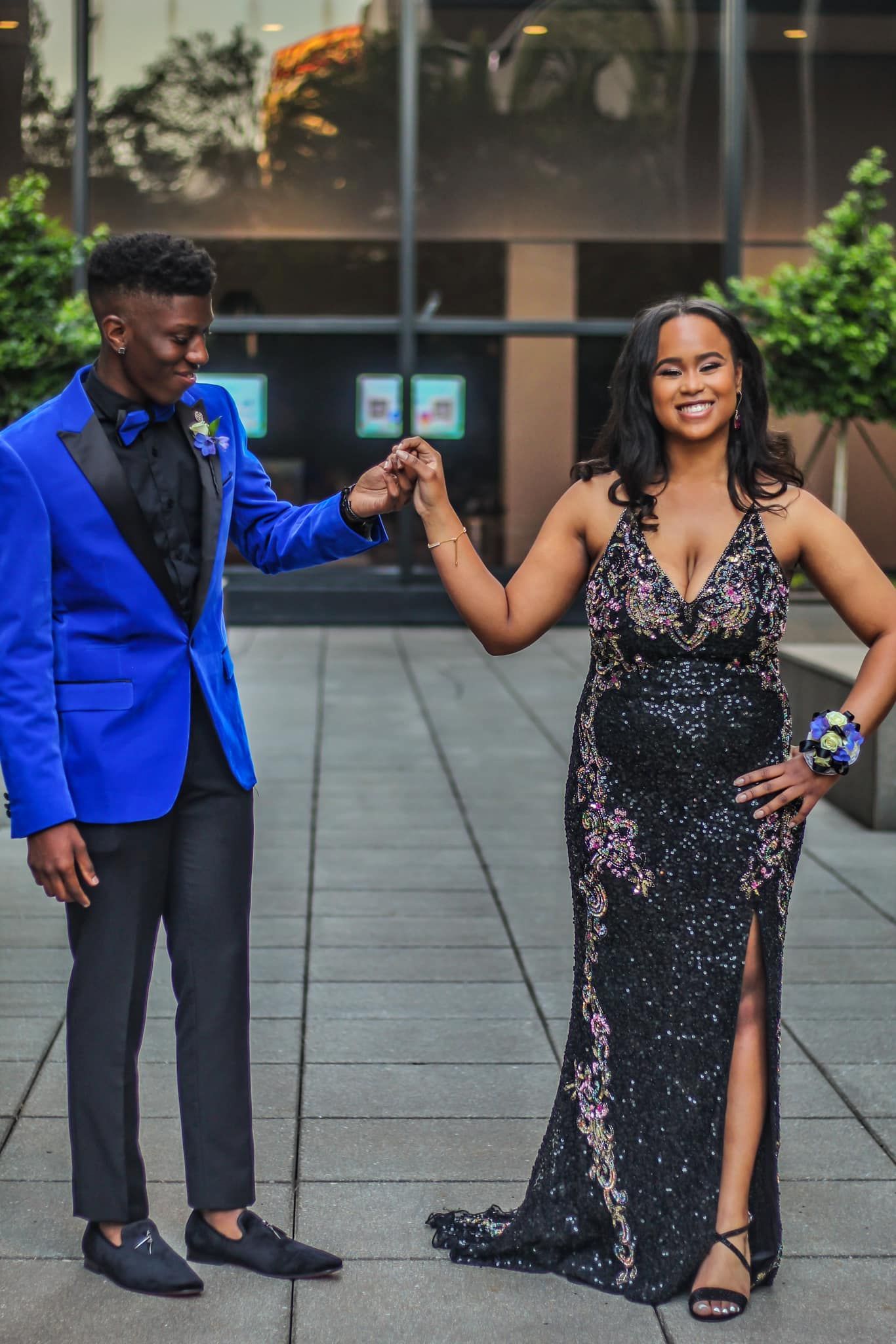 A smiling prom couple poses outdoors, the man in a royal blue tuxedo and woman in a black gown.