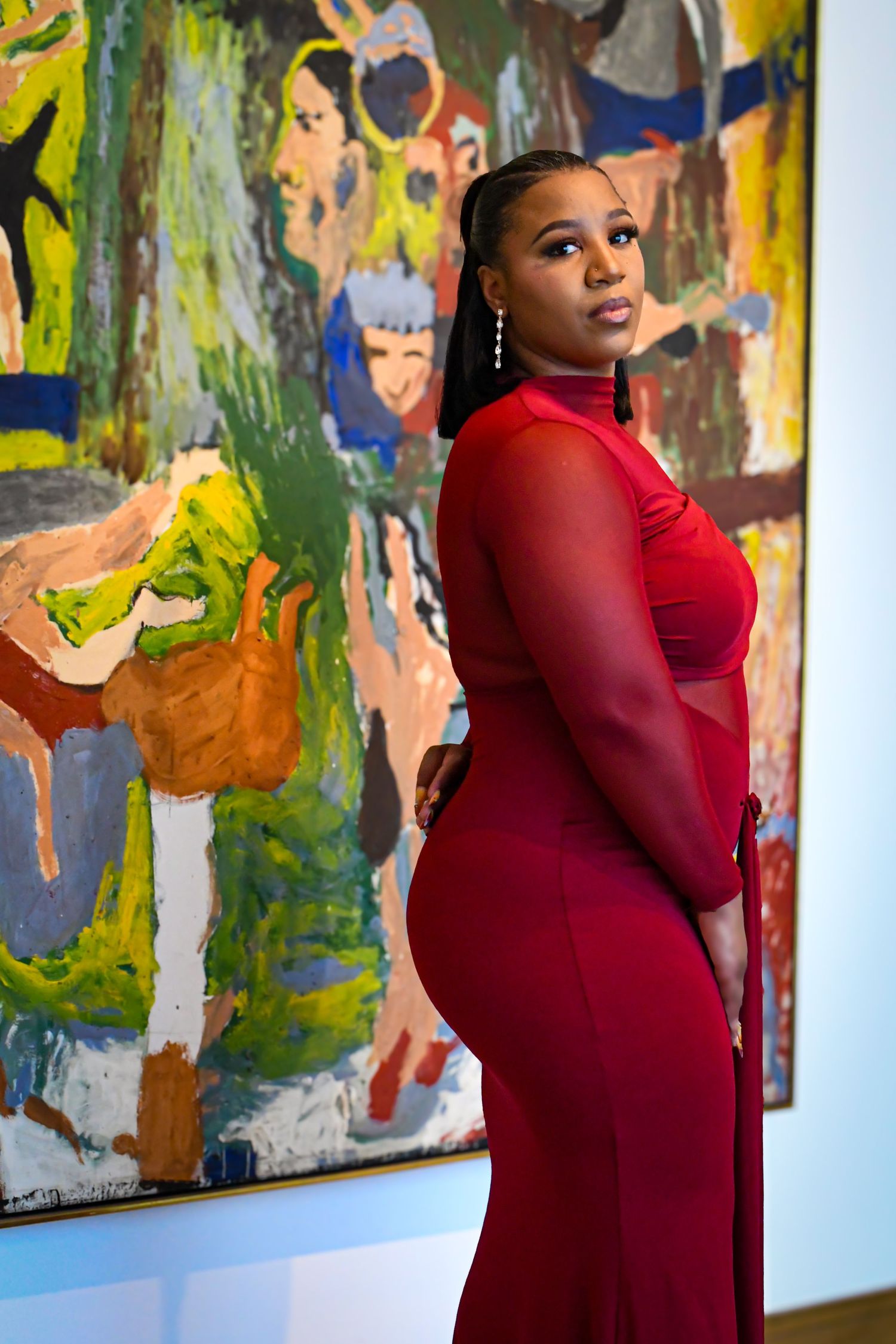 Woman in red gown poses in front of colorful abstract painting in art gallery.