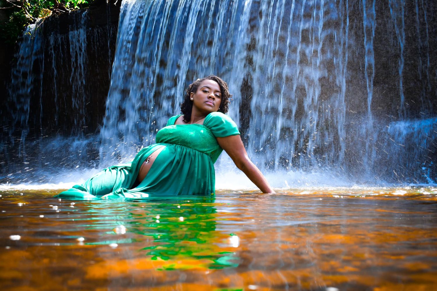 A pregnant woman in a flowing teal dress sits gracefully in a shallow pool before a cascading waterfall.
