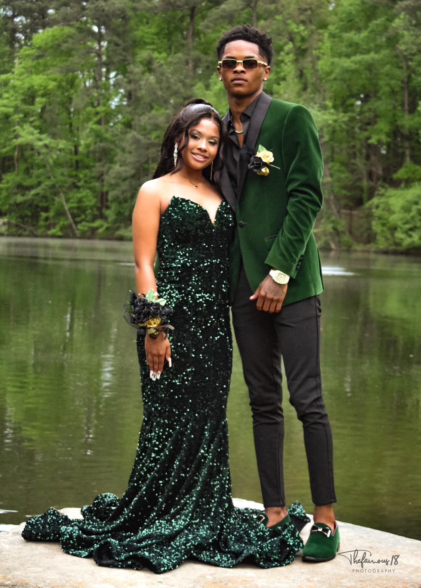 A couple dressed in matching emerald green sequin prom outfits poses by a serene forest lake.