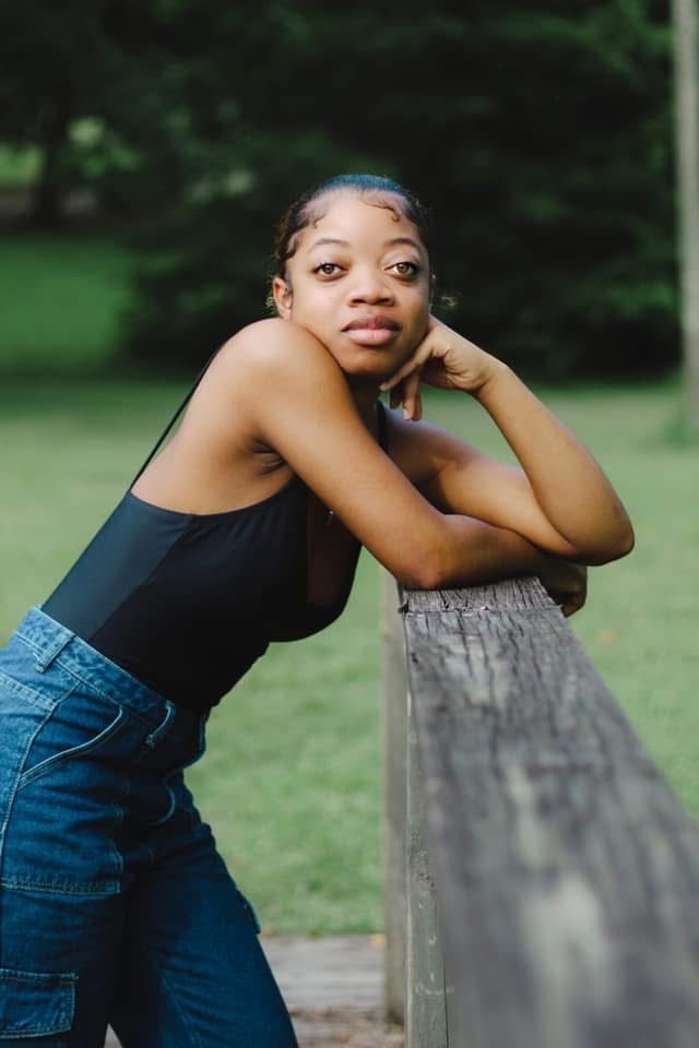 Young Black woman in black top and jeans leaning on wooden fence in park, portrait photography.