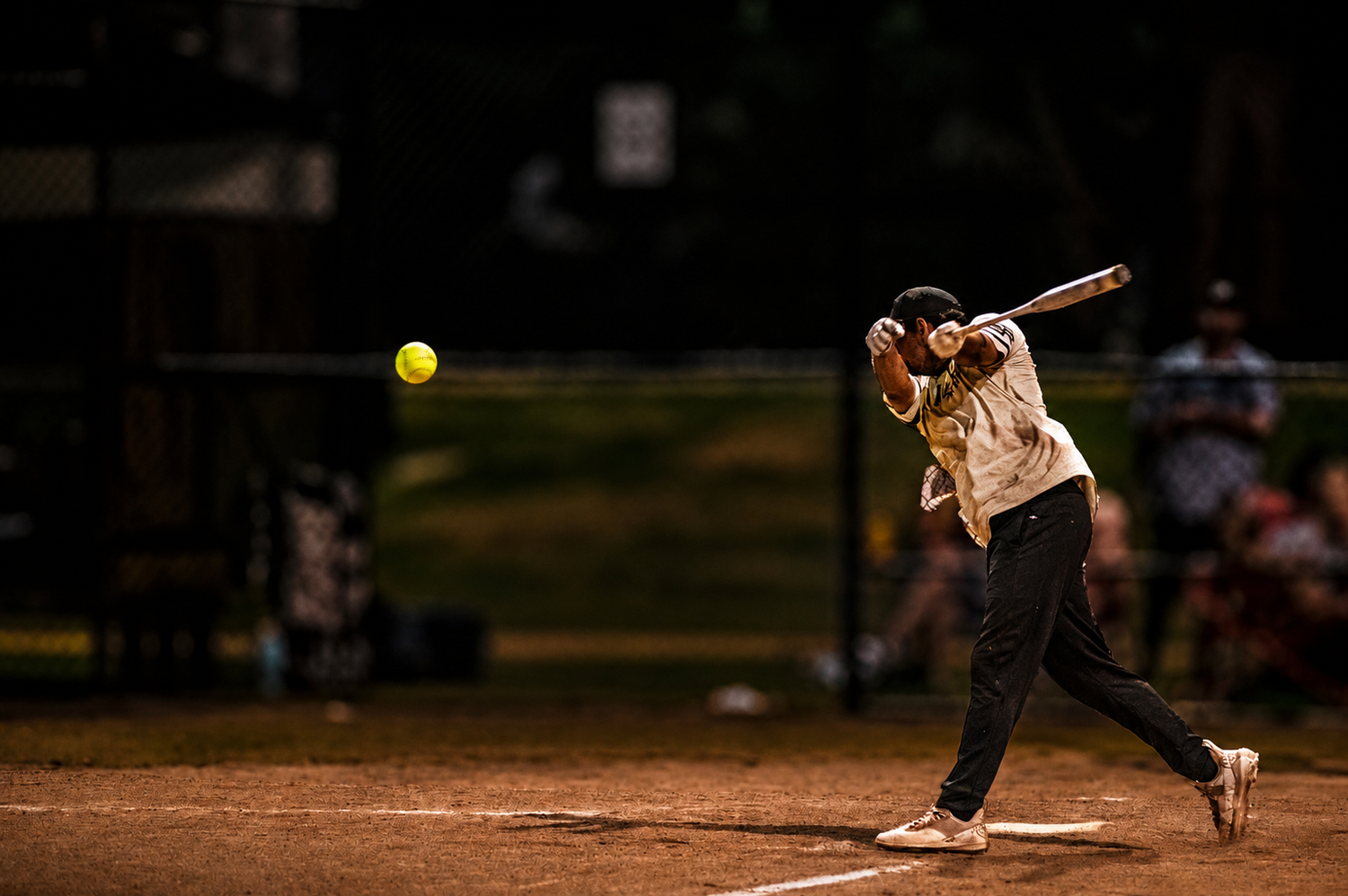Softball batter swinging at a yellow ball during a game on a dirt field under dark outdoor lighting.