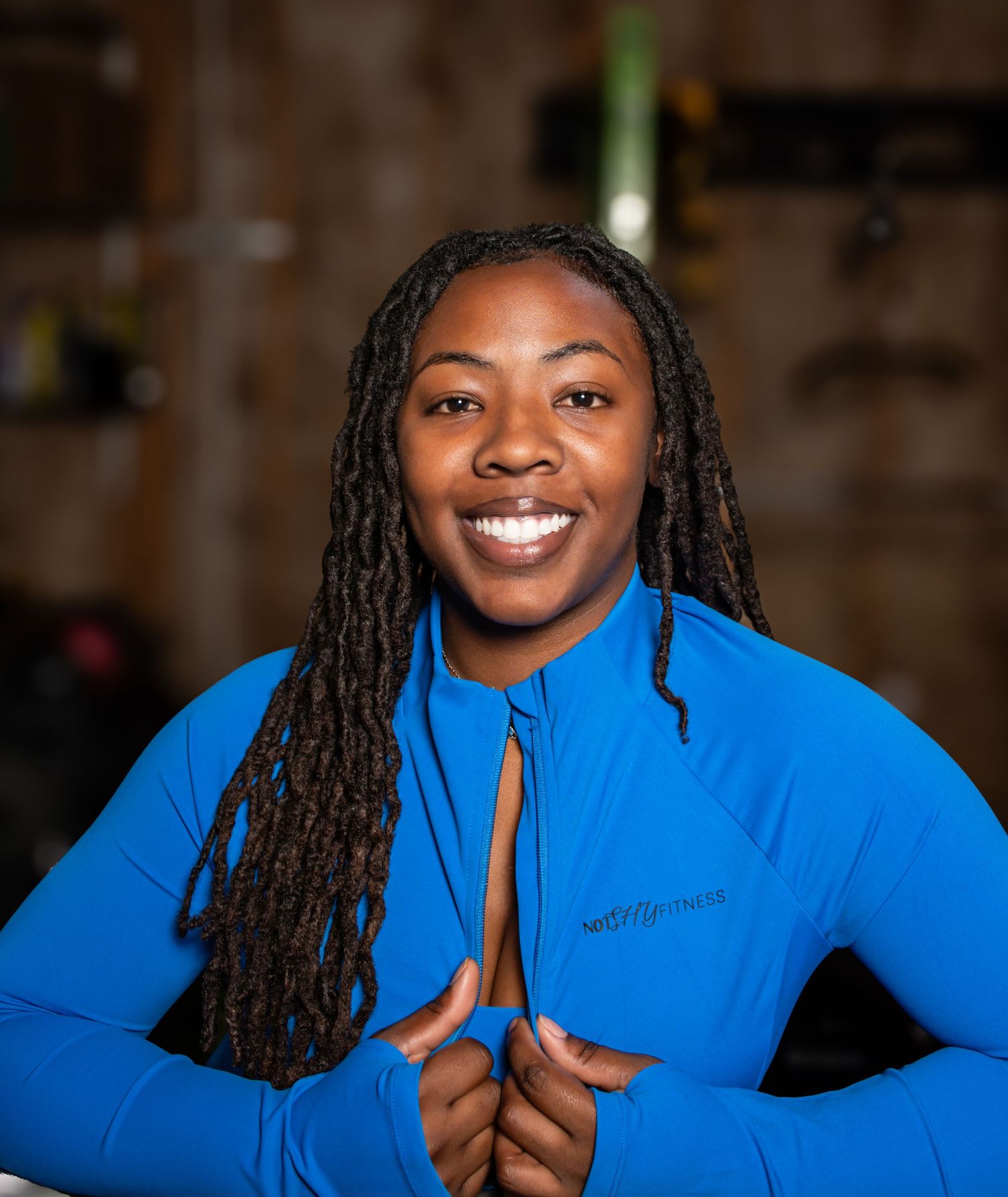 A smiling female fitness trainer in a blue zip-up athletic top poses confidently in a gym setting.