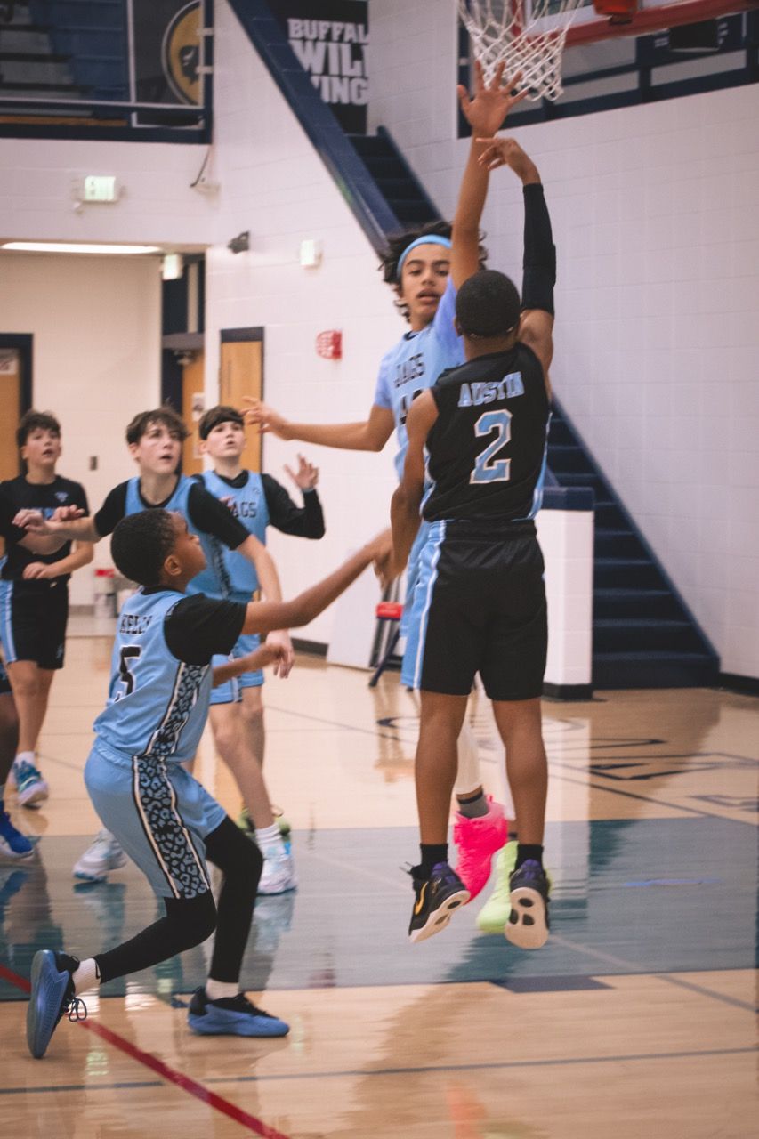 A youth basketball player in black jersey number 2 leaps to block a shot near the hoop.