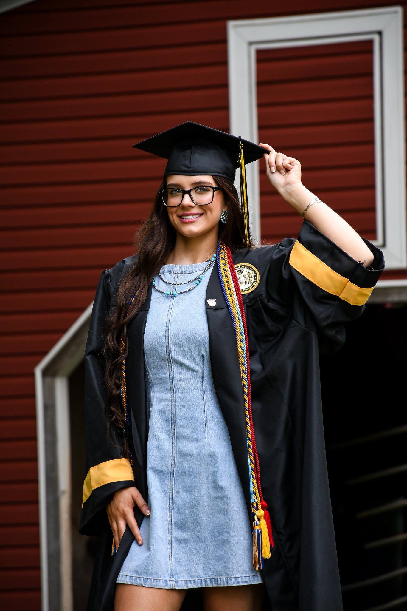 A smiling female graduate in cap and gown with gold trim and honor cords poses proudly outside a red building.