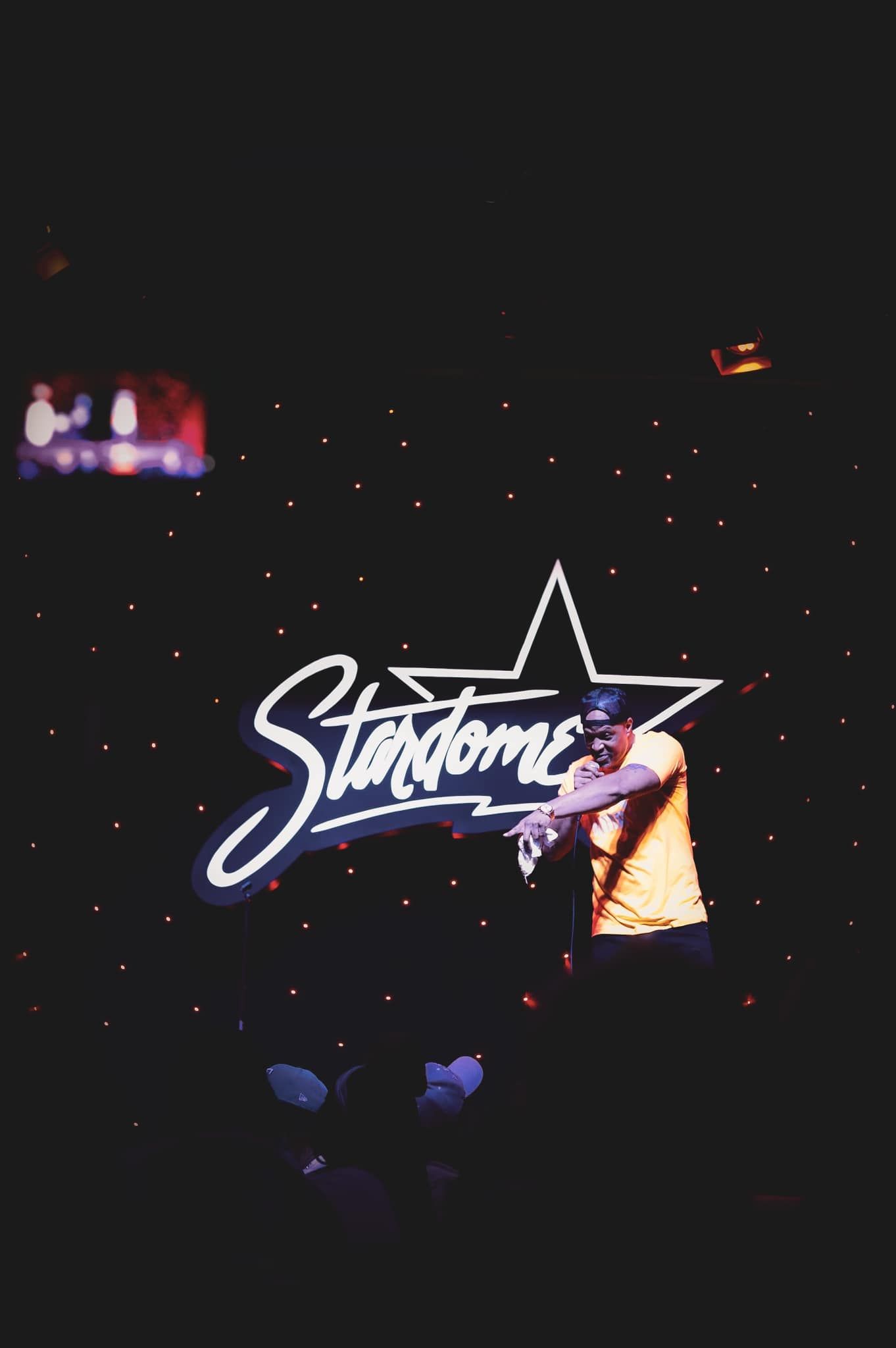 A performer on stage at the Stardome comedy club illuminated by a glowing neon star logo sign.
